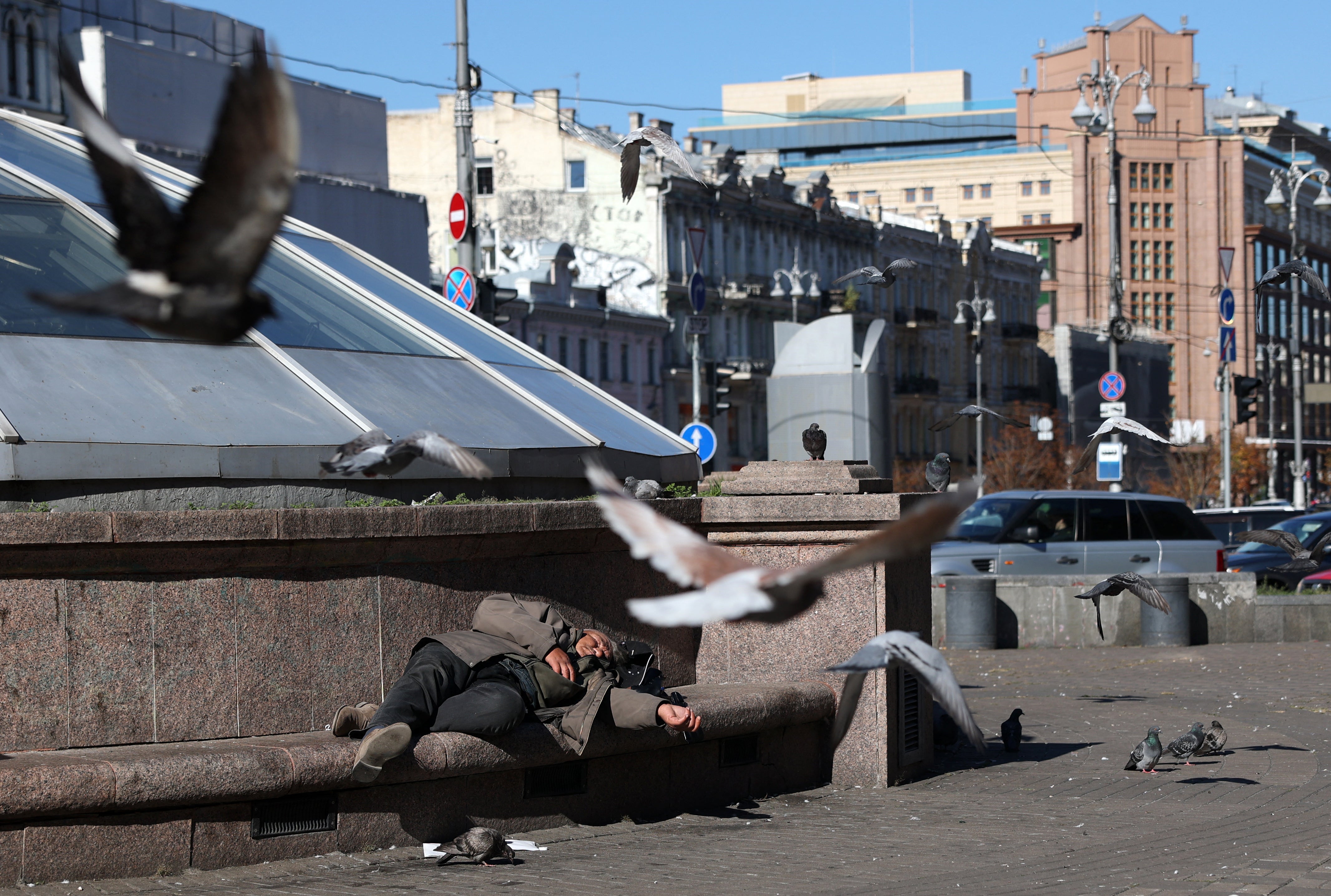 An elderly man lays on a bench as pigeons fly past on a sunny autumn day in central Kyiv